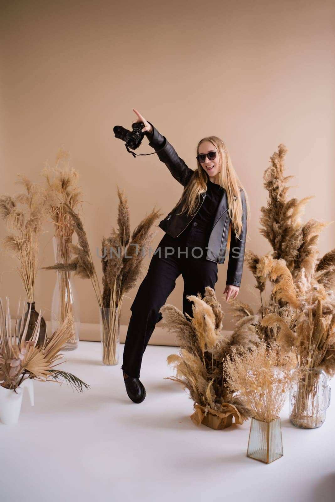 A crazy woman photographer, a blonde with a camera, happy and smiling in the production studio. Wearing a black clothes on a brown background