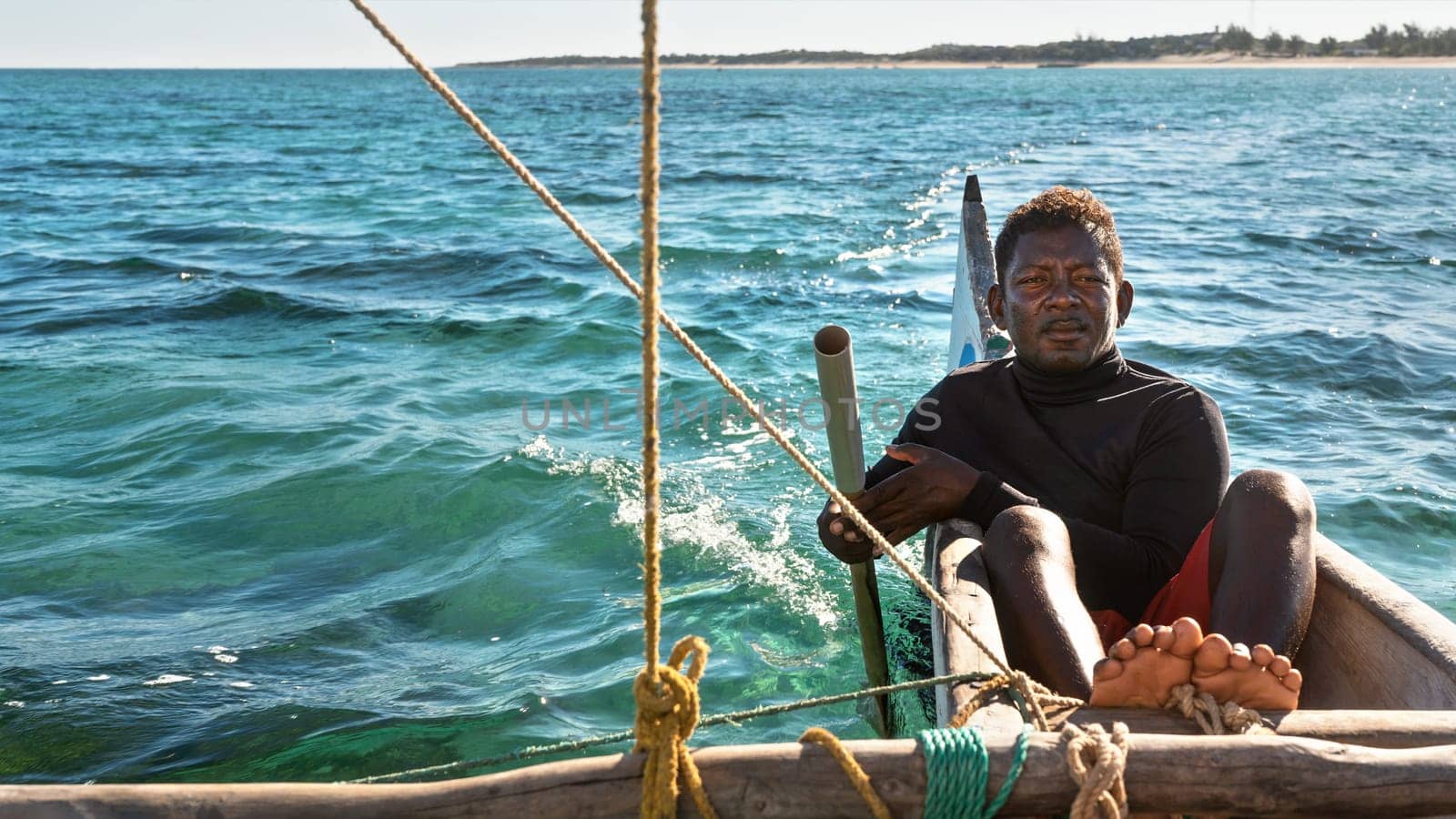 Anakao, Madagascar - May 03, 2019: Unknown Malagasy fisherman sitting on back of his piroga (small fishing boat), leading direction with simple rudder, green blue sea during sunny day in background by Ivanko