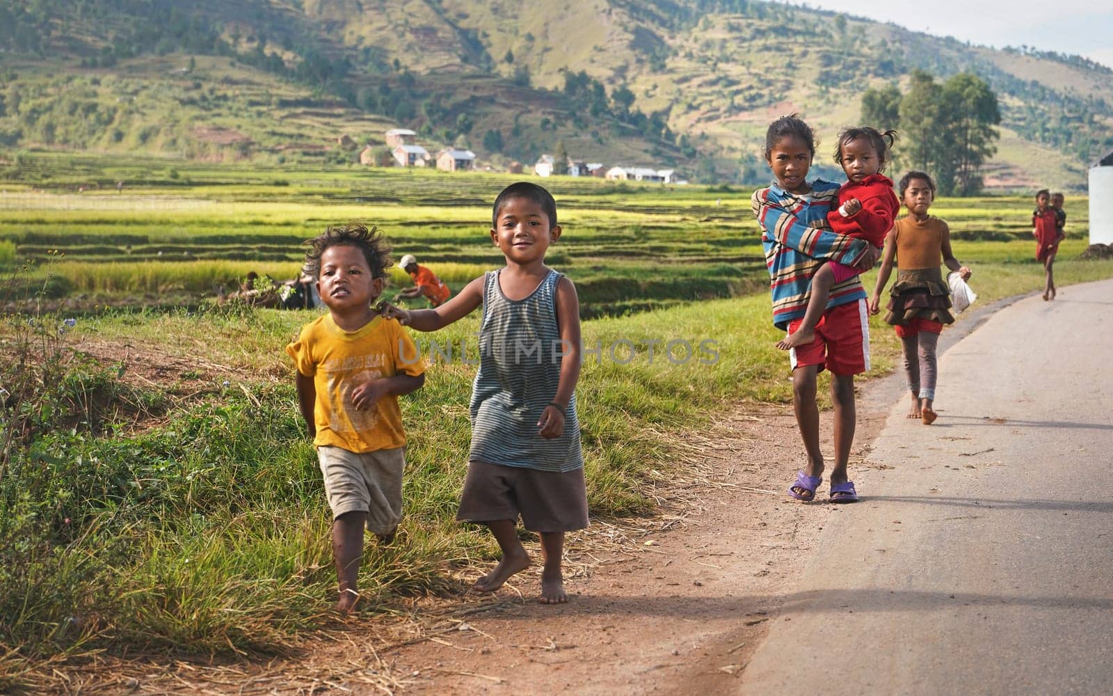 Manandoana, Madagascar - April 26, 2019: Group of unknown Malagasy kids running on road next to rice field, small hills in background, people at Madagascar are poor but cheerful, especially children