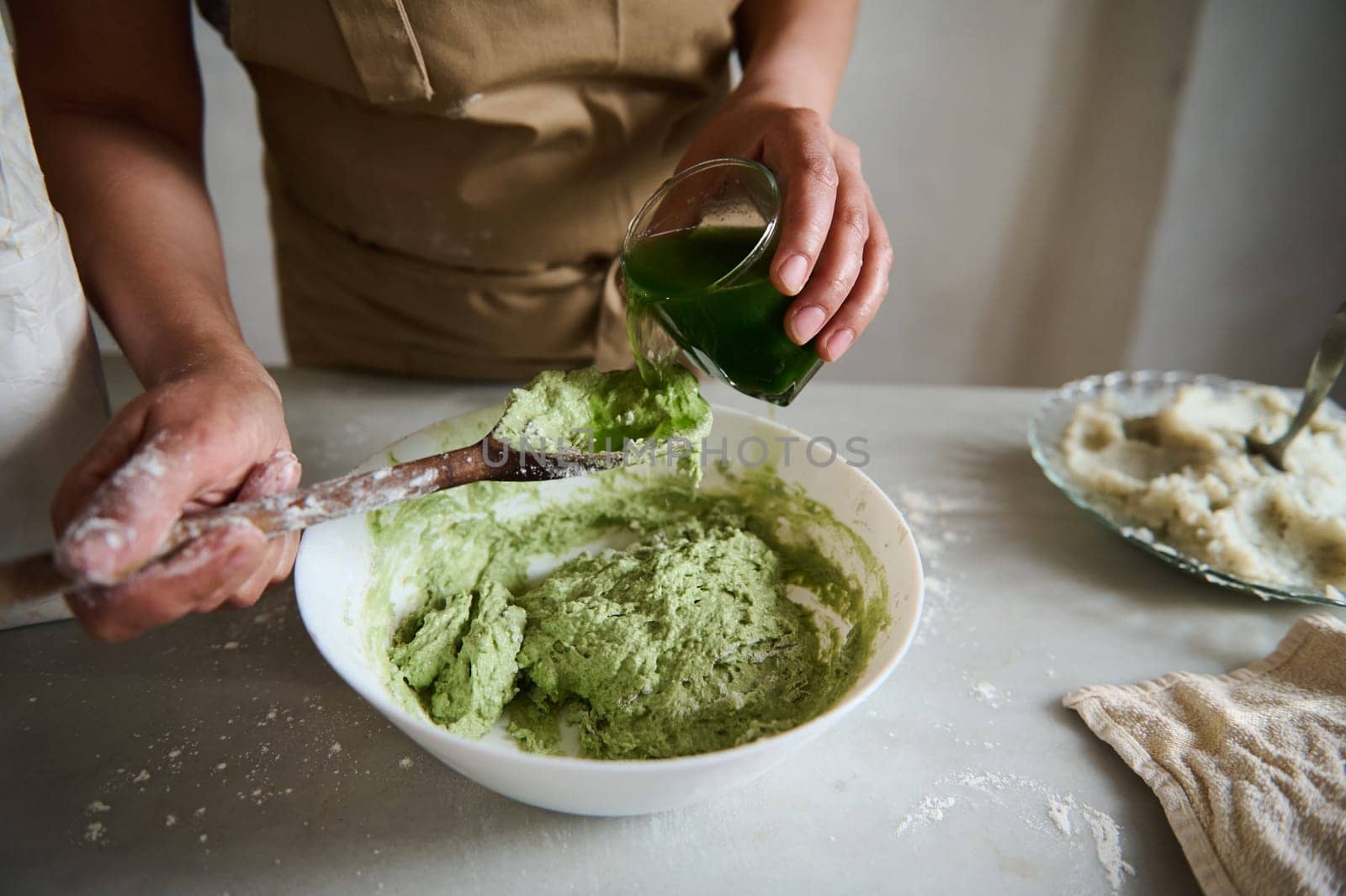 Close-up hands of a chef pastry, housewife pouring green spinach water into a bowl with wheat four, making dough of green color for ravioli or dumplings with mashed potato, in the rustic home kitchen