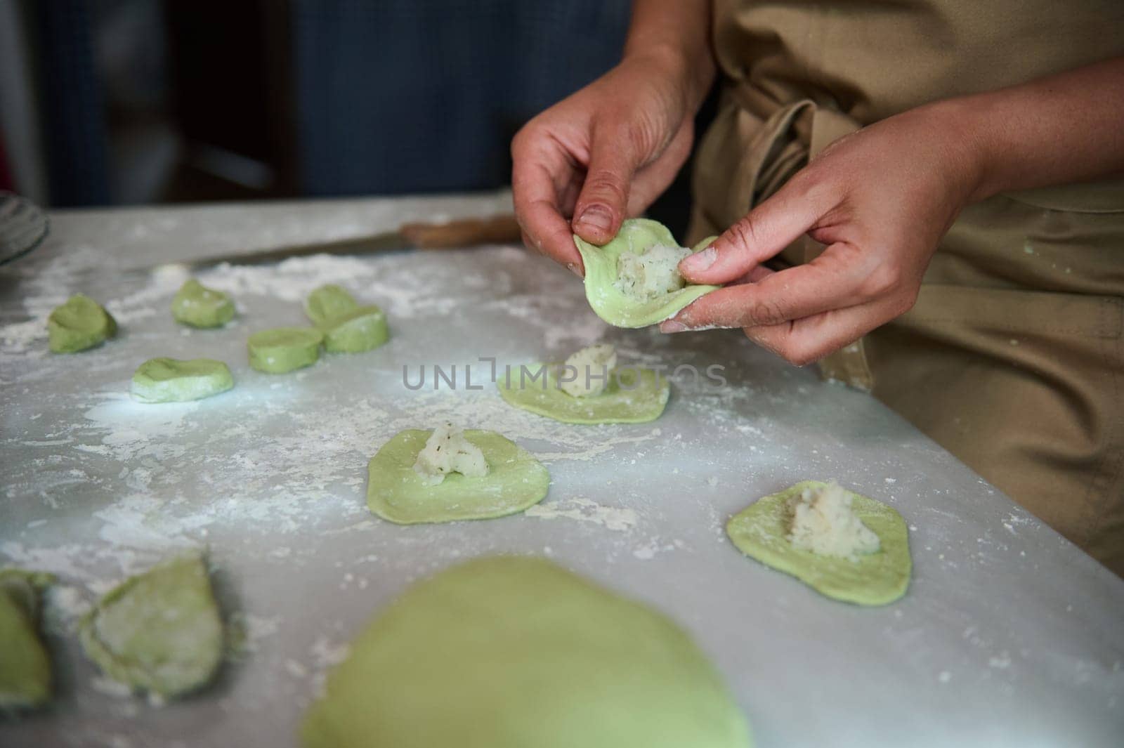Close-up woman stuffing dough with mashed potatoes, modeling dumplings, standing at kitchen table with kneaded dough and ingredients. The process of cooking ravioli, varenniki, vegetarian pelmeni by artgf