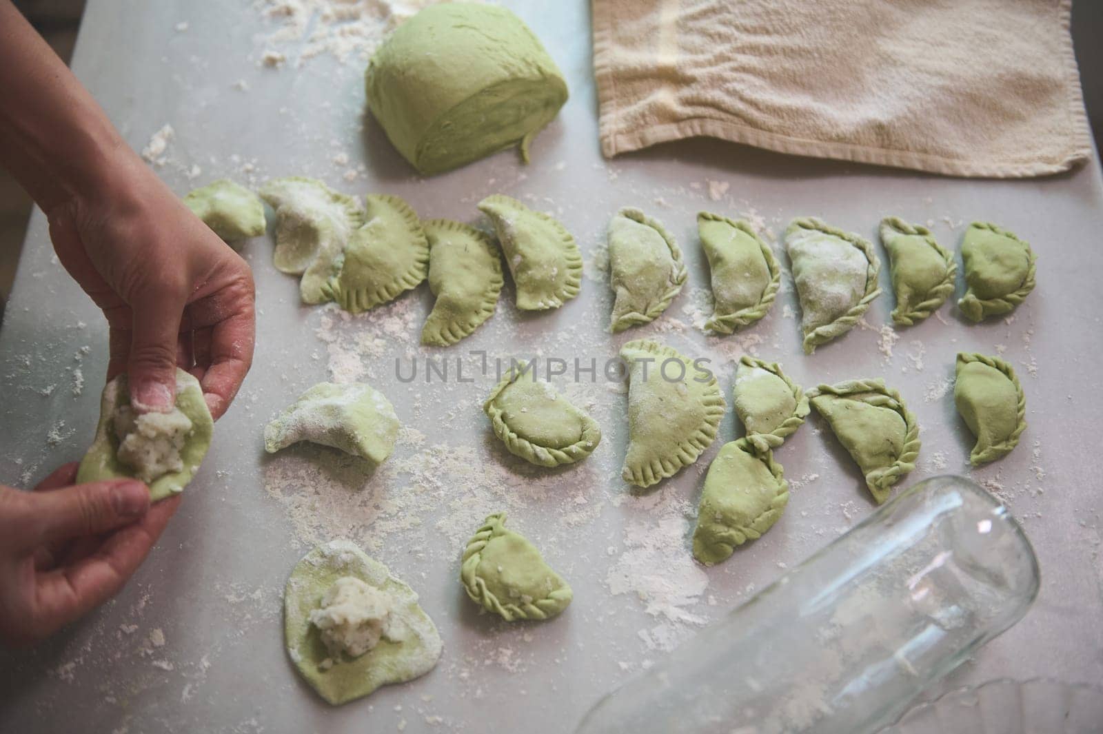 Details on the hands of a woman housewife, chef in beige apron making dumplings from green dough with melted spinach, and filling it with mashed potatoes, standing at kitchen table in rural house