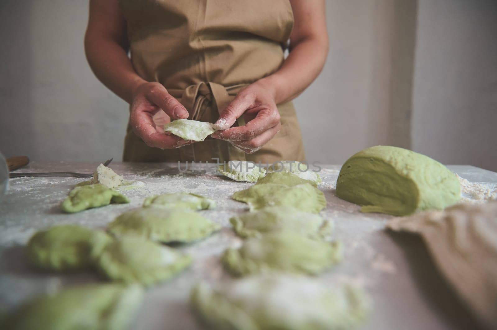 Housewife filling rolled dough with mashed potatoes, preparing homemade varenyky, dumplings, ravioli according to traditional family recipe, standing at marble kitchen table with ingredients. Top view