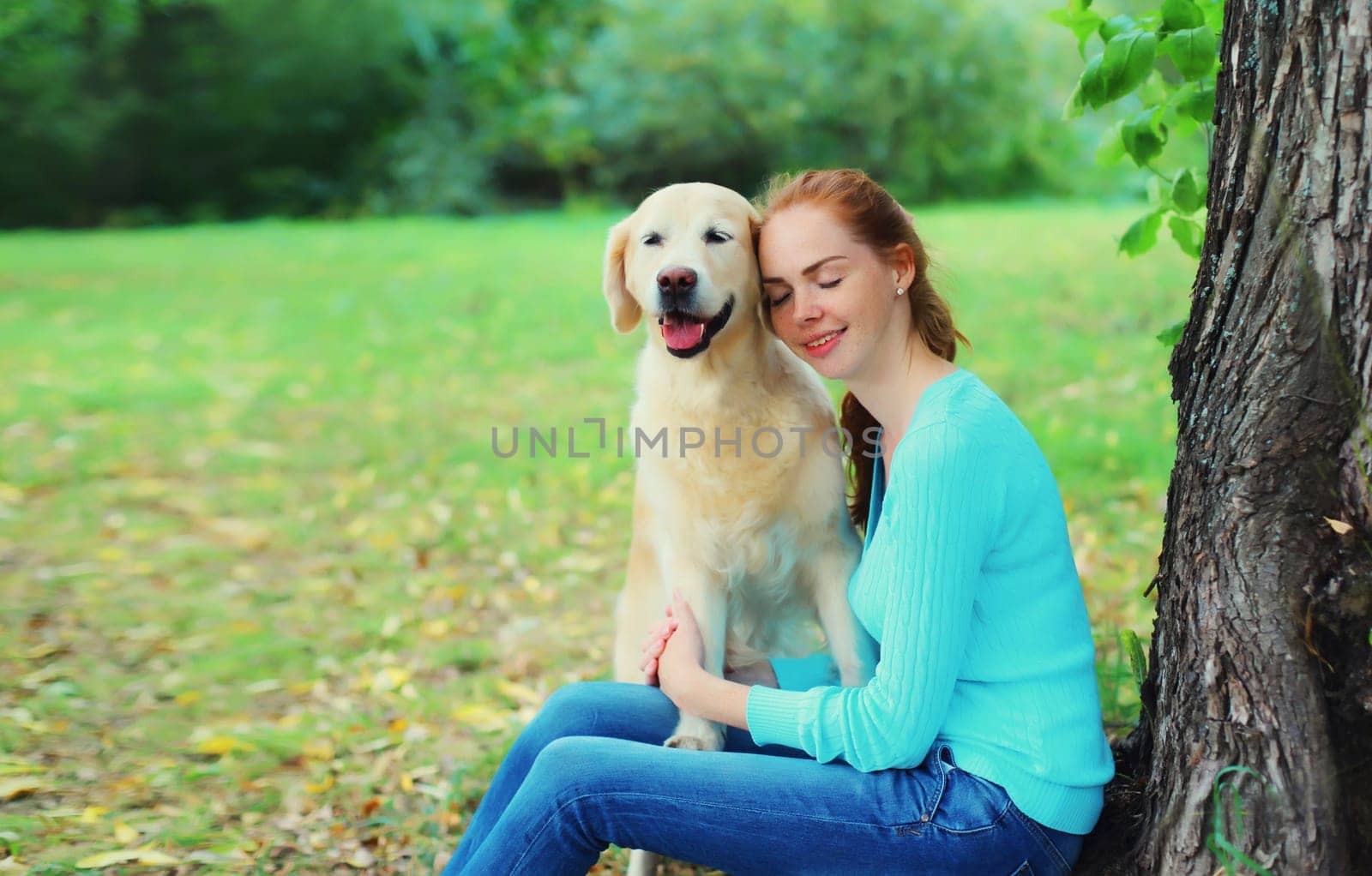 Happy owner young woman with Golden Retriever dog sitting together under tree in summer park