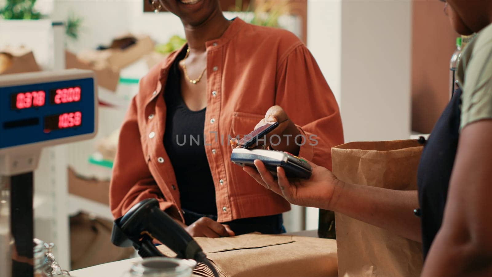 Woman using mobile nfc payment to pay for bio produce from local farm, supporting sustainable lifestyle and small business. African american merchant and buyer chatting at cash register.