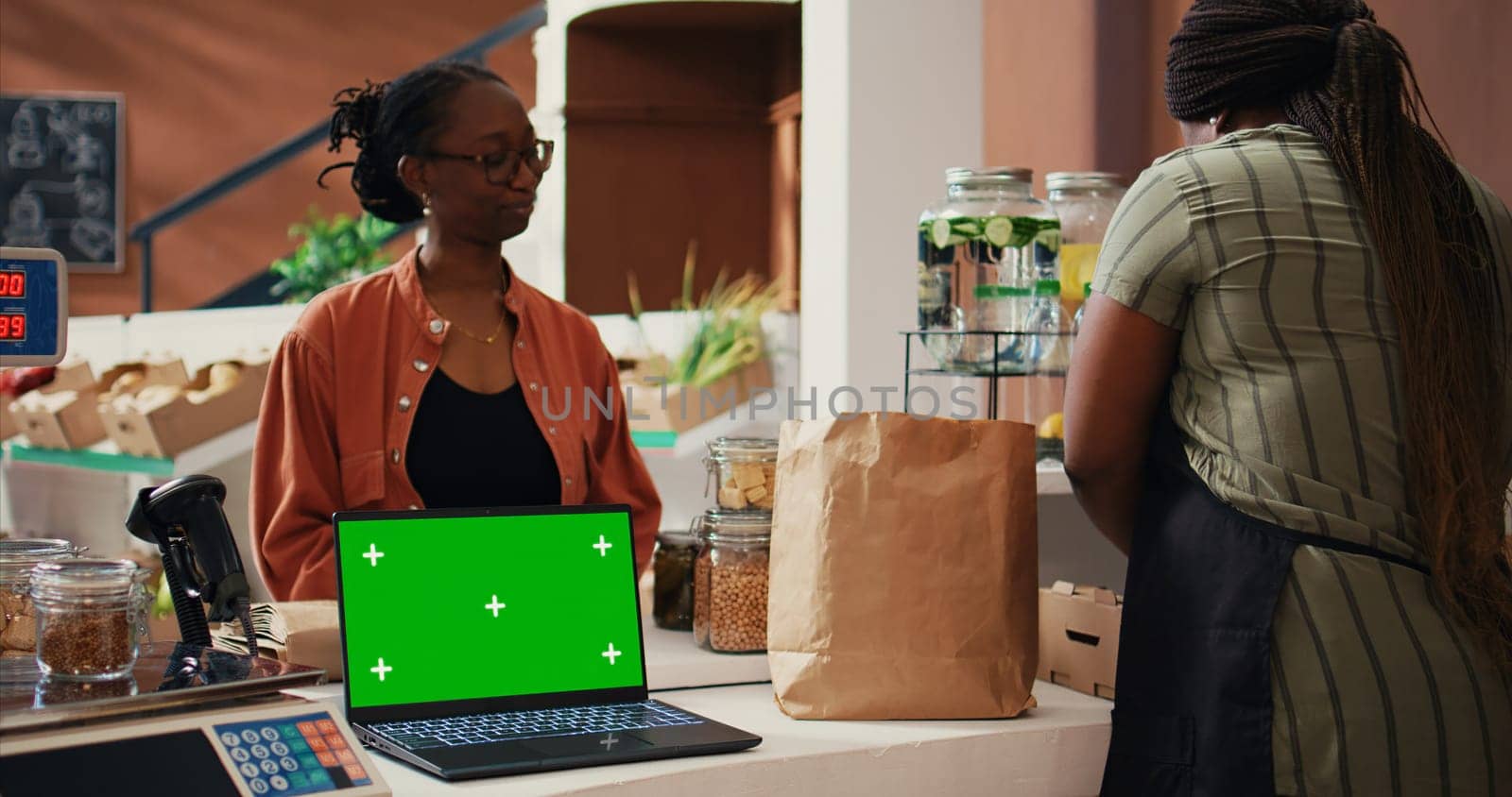 Retailer having laptop that shows greenscreen on display at cash register, giving eco products to client at market. Woman using computer with isolated chromakey template, mockup layout. Tripod shot.