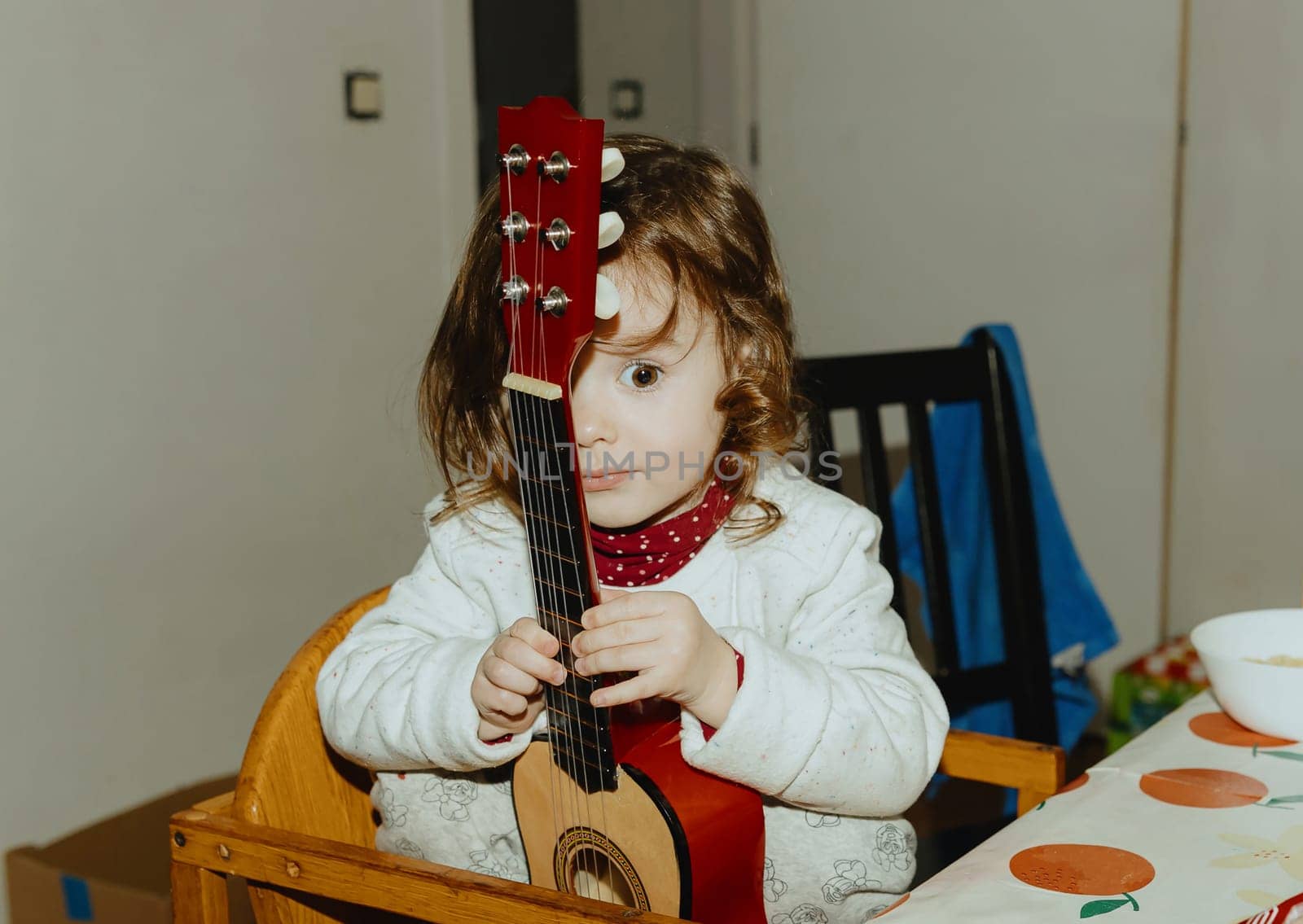 One beautiful Caucasian baby girl holds a guitar with one eye closed, sitting on a wooden chair at the table in the kitchen, side view close-up.
