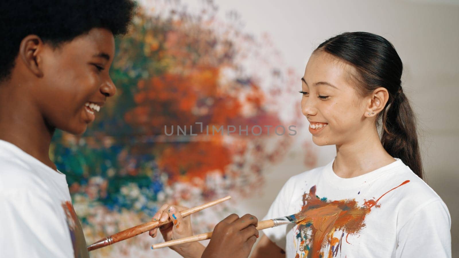 Smiling diverse children using paint brush painted color on each other white shirt shirt at colorful stained wall in art lesson. Represent exchanging experience, learning each other. Edification.