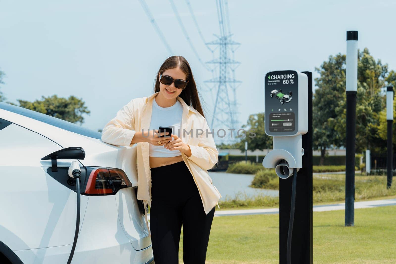 Young woman use smartphone to pay for electricity at public EV car charging station green city park. Modern environmental and sustainable urban lifestyle with EV vehicle. Expedient