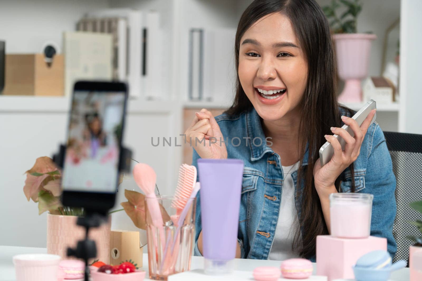 Young beautiful Asian reviewing cosmetic makeup and skin care collection, talking customer on social media online, presenting product surrounded beauty merchandise recording blurred phone . Stratagem.