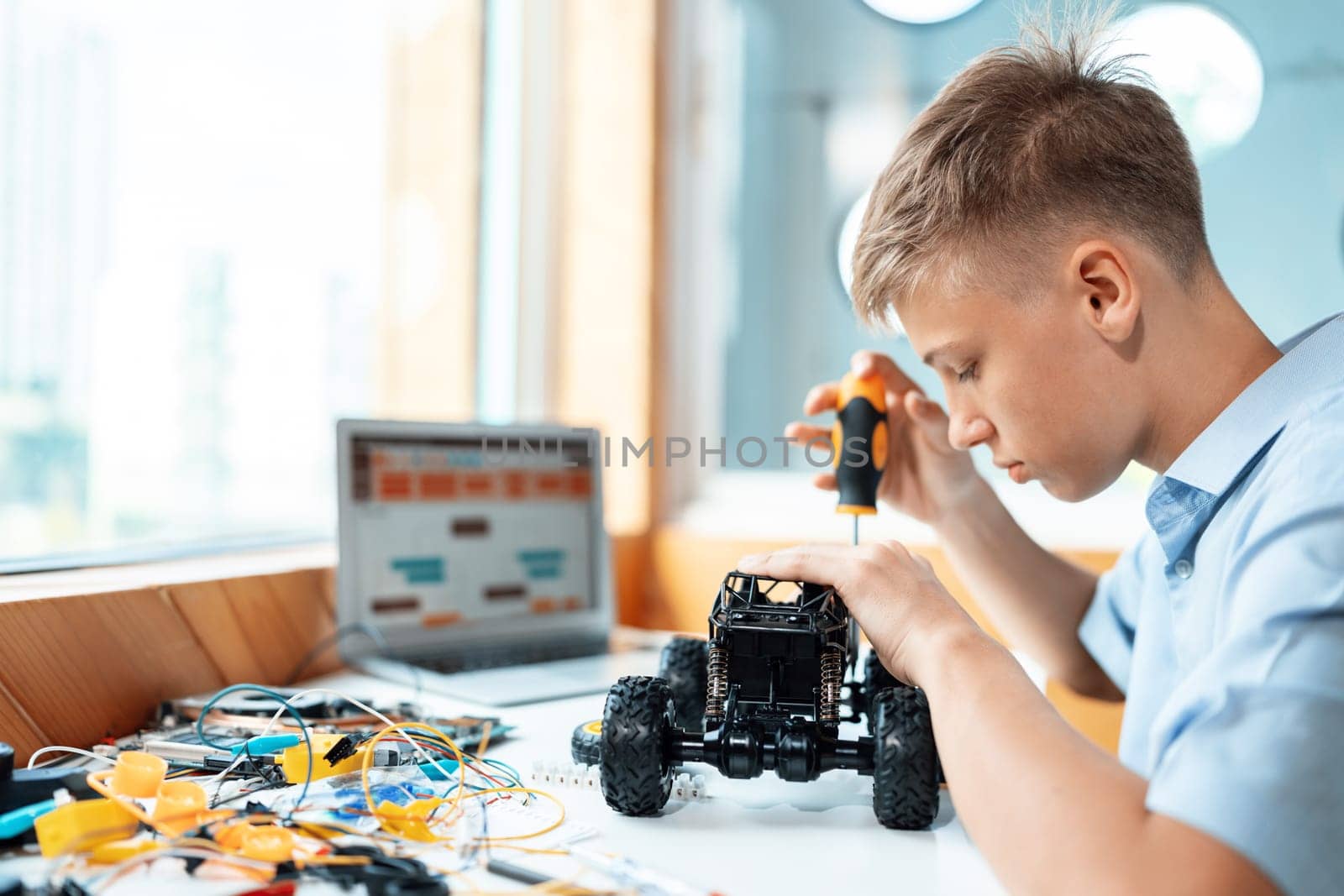 Blonde hair schoolboy using screwdriver fixing robotics vehicle learning technology in STEM class. On table put laptop, controller, electric wire, battery charger, and robotic vehicle. Edification.