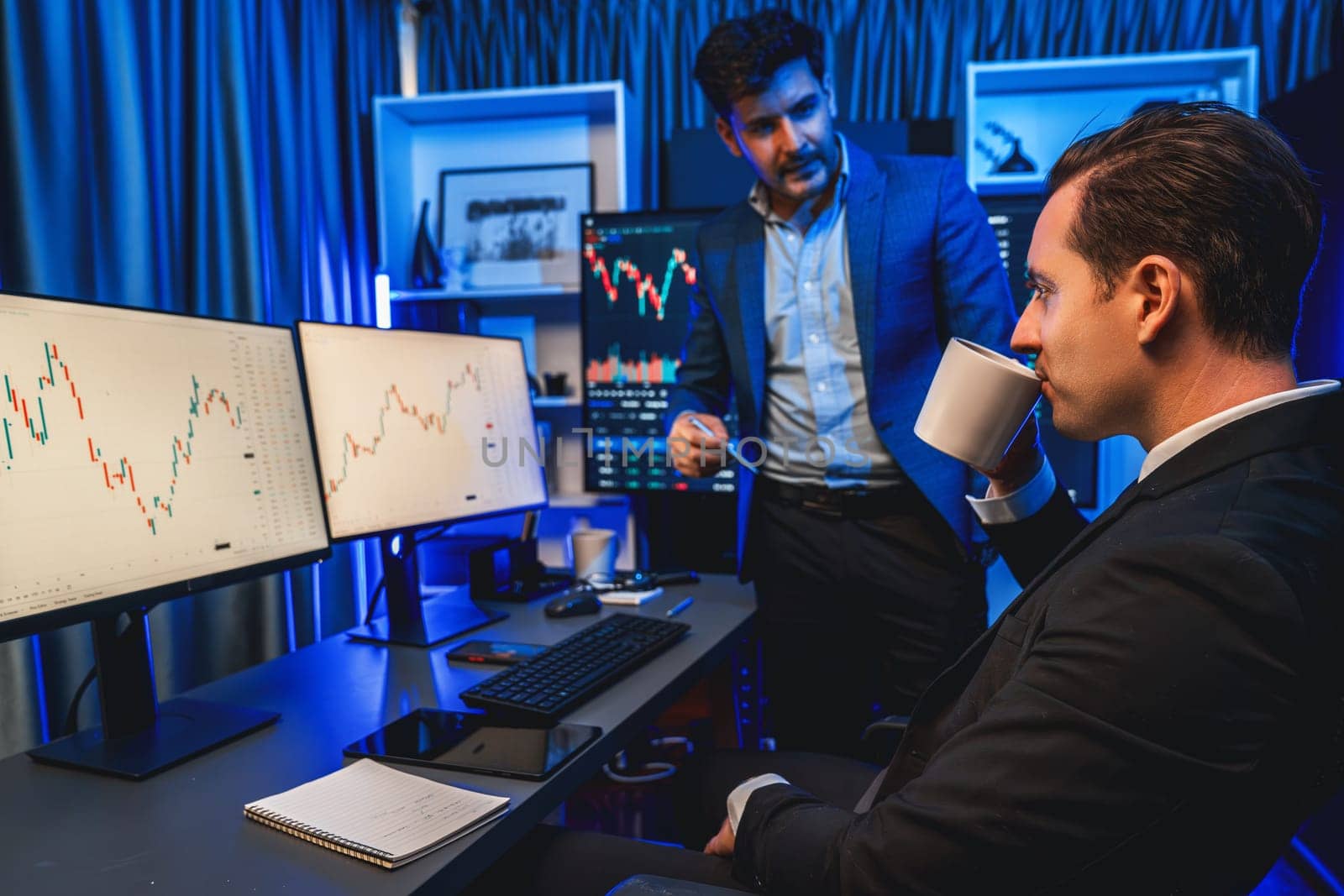 Two stock exchange traders discussing dynamic investment graph in currency rate on monitor at night. Businessman partners coffee meeting in high stock market in neon light at workplace. Sellable.