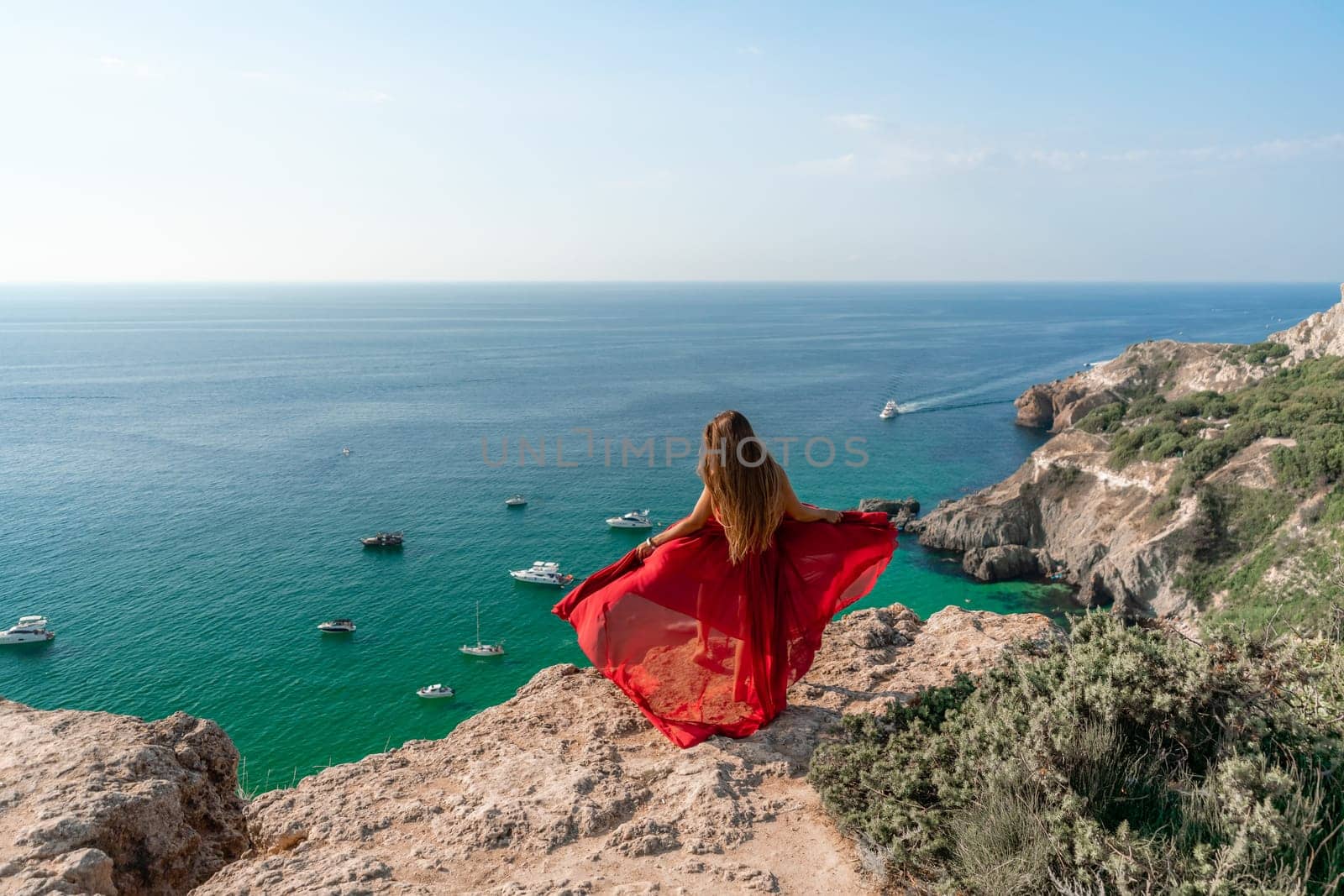 Woman sea red dress yachts. A beautiful woman in a red dress poses on a cliff overlooking the sea on a sunny day. Boats and yachts dot the background