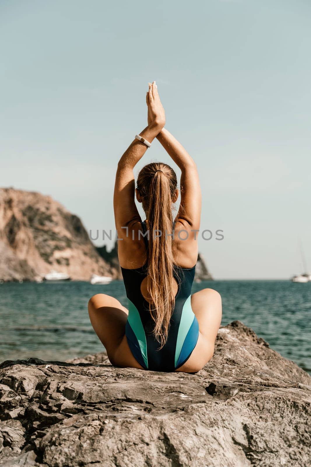 Yoga on the beach. A happy woman meditating in a yoga pose on the beach, surrounded by the ocean and rock mountains, promoting a healthy lifestyle outdoors in nature, and inspiring fitness concept