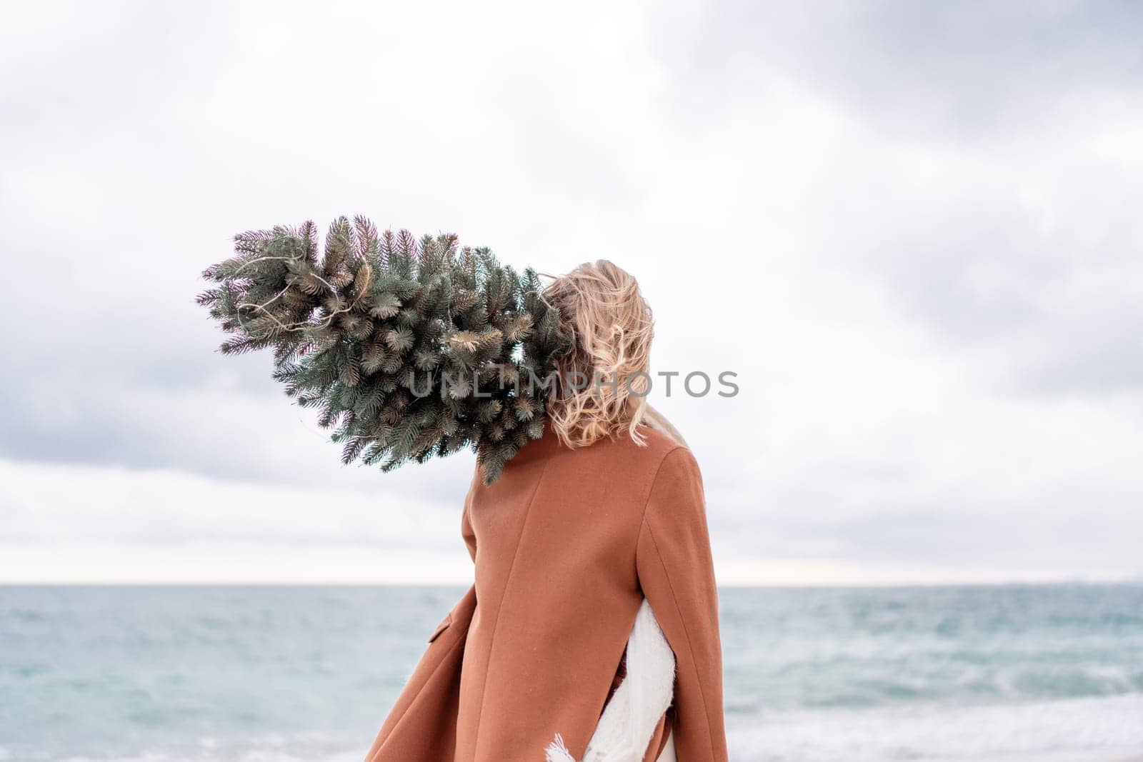 Blond woman Christmas tree sea. Christmas portrait of a happy woman walking along the beach and holding a Christmas tree on her shoulder. She is wearing a brown coat and a white suit. by Matiunina