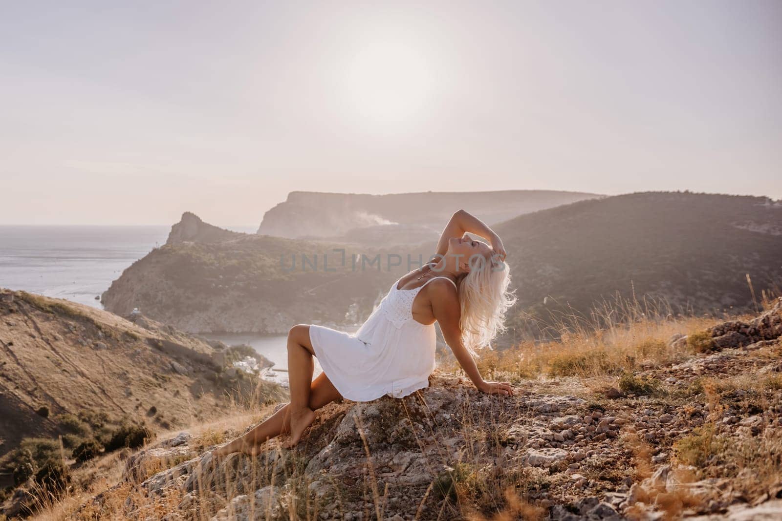 A woman is sitting on a hillside overlooking the ocean. She is wearing a white dress and has blonde hair. The scene is serene and peaceful, with the ocean in the background. by Matiunina