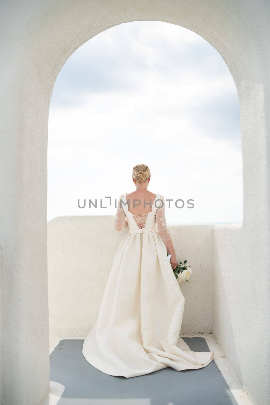 A bride stands in front of an archway, holding a bouquet of flowers. The archway is white and the sky is cloudy. The bride is wearing a white dress and her hair is styled in a bun. by Matiunina