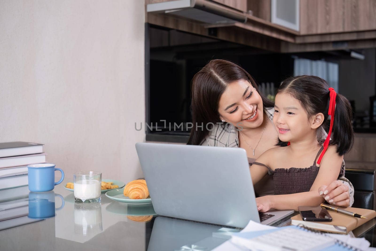 a single mother, sits at home working on a laptop with her daughter beside her watching and encouraging her..