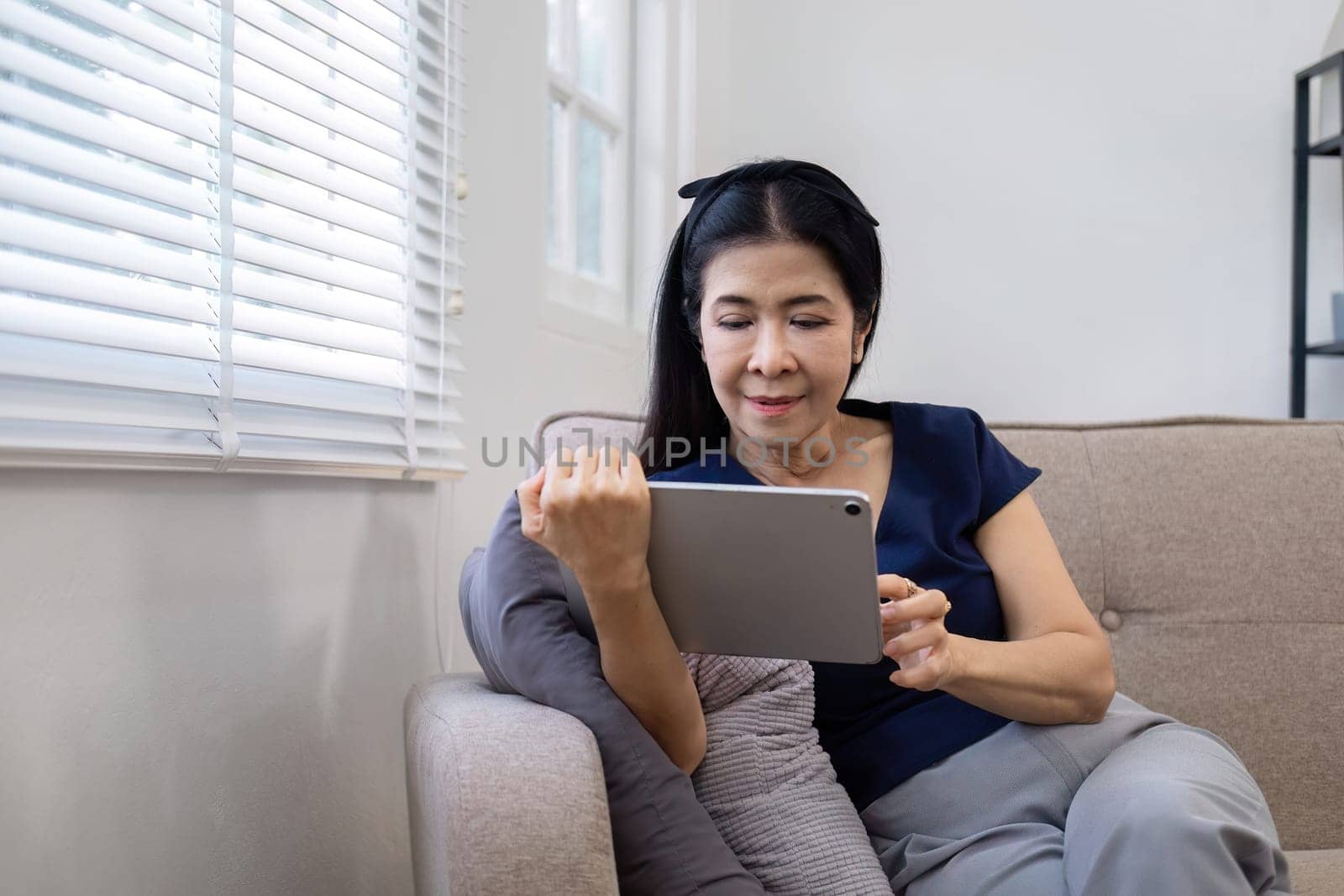 Elderly woman's leisure day watching social media on tablet, watching news on sofa in living room by wichayada