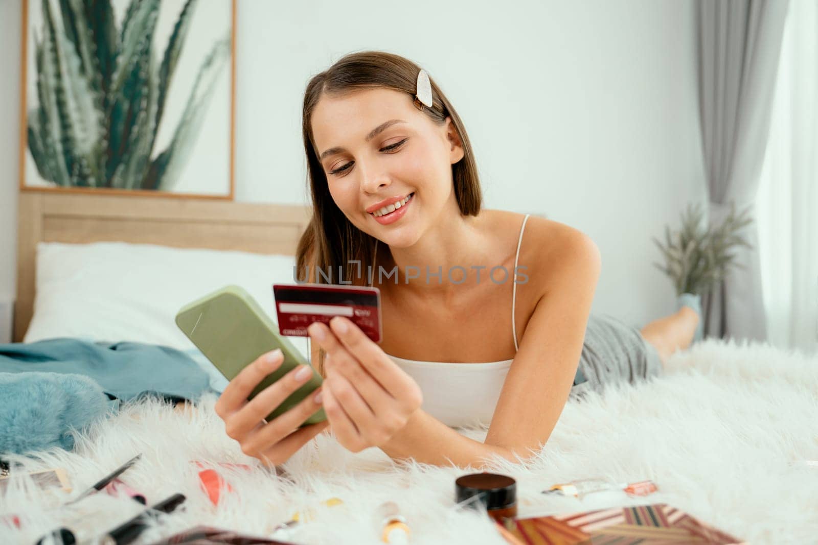 Young woman lying on the bed using online payment app and digital wallet on smartphone to pay with credit card. E commerce shopping and modern purchasing via mobile internet. Unveiling