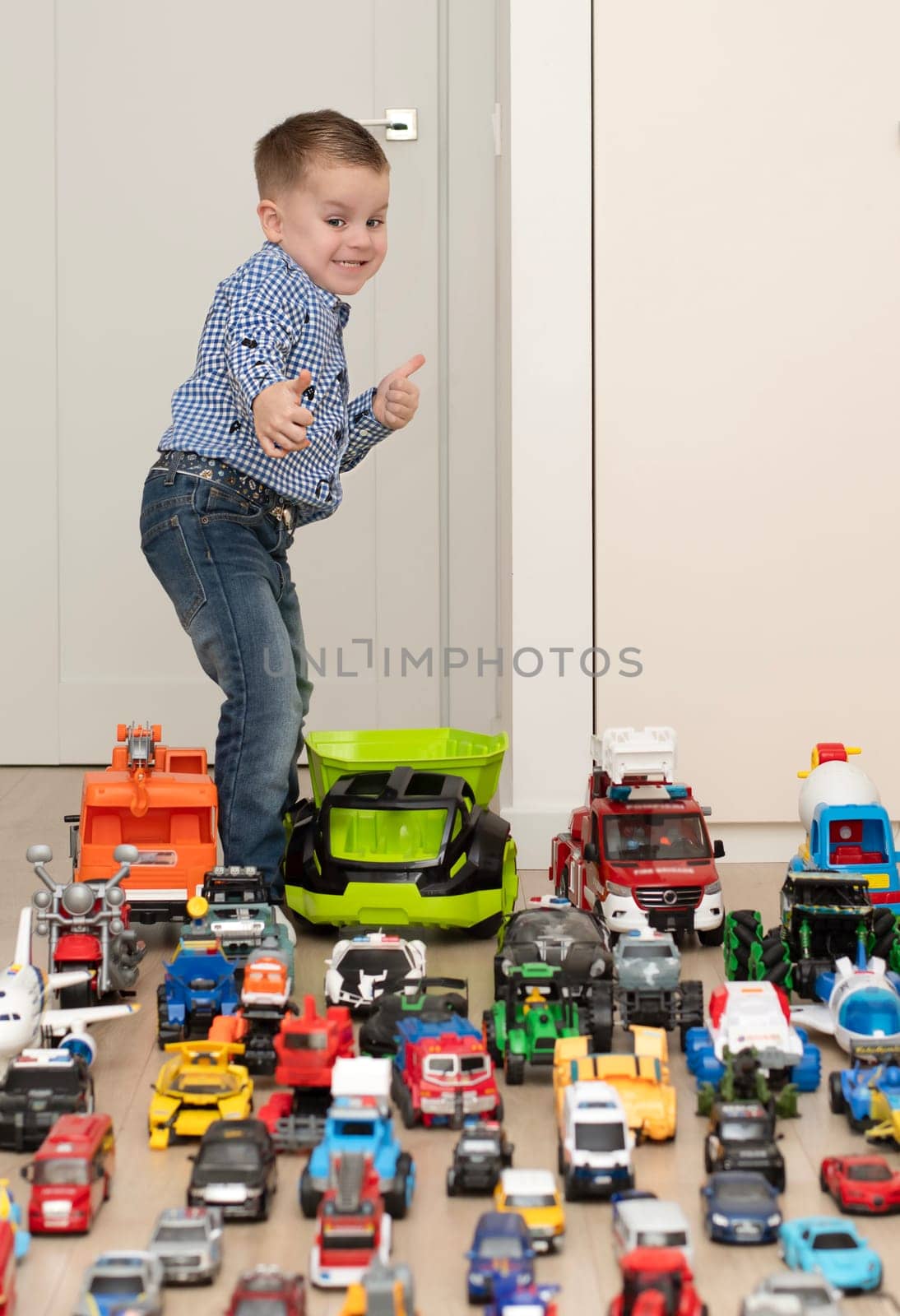 Children. Toy cars. A small cheerful and handsome boy, 4 years old, wearing a checkered shirt, plays with many colorful cars in a home interior. The toys are placed evenly on the floor.