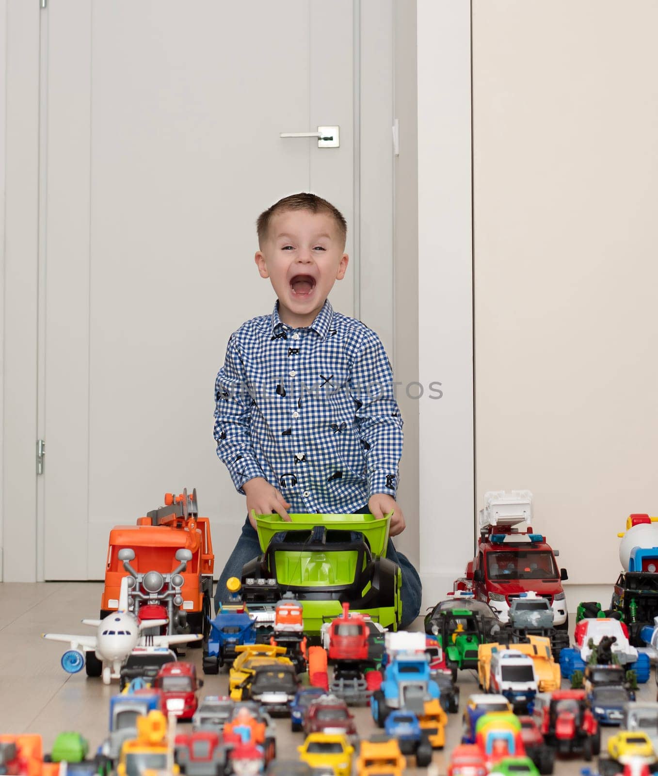 Concept of children's toys. A little boy, 4 years old, cheerful and handsome, happily plays, sitting on the floor, with multi-colored small and large cars in the children's room. Soft focus