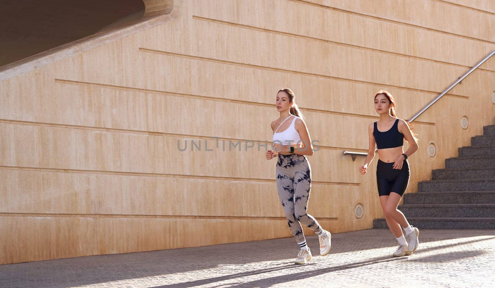 Two active women athlete running on sunny morning side by side on modern buildings background