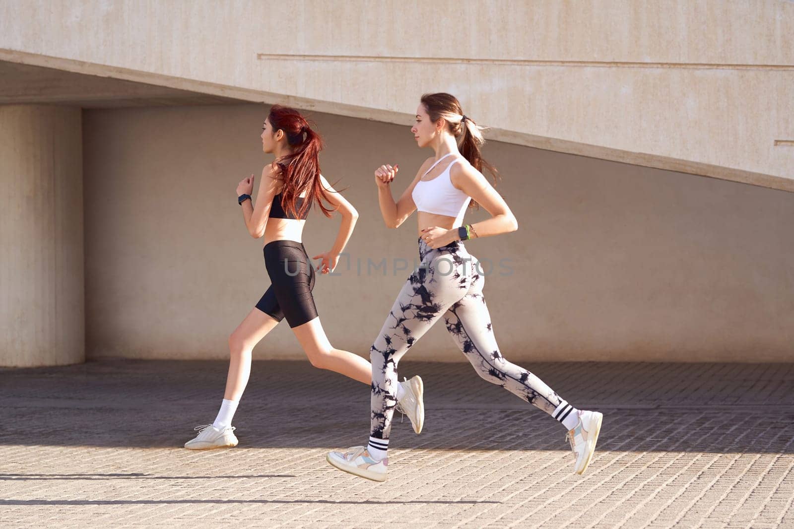 Two young women in sportswear are running on modern building background. Active lifestyle concept