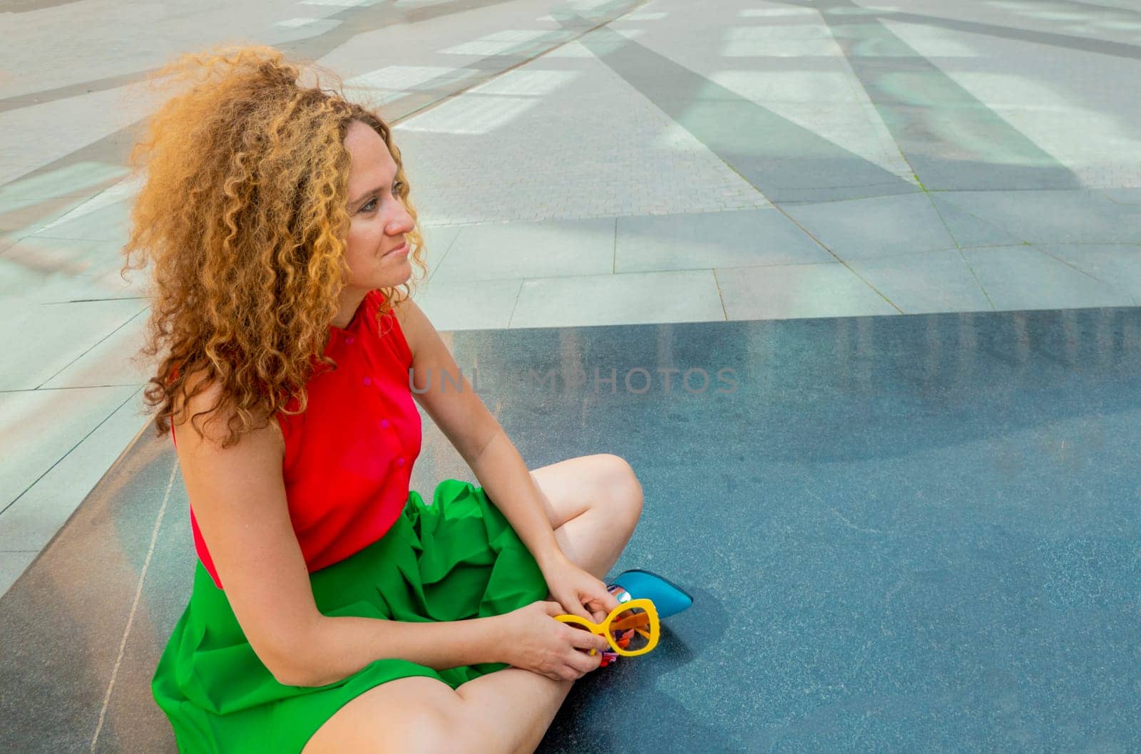 a fashionable woman in bright clothes with long blond curly hair is sitting in the lotus position on the street basking in the sun and resting by kajasja
