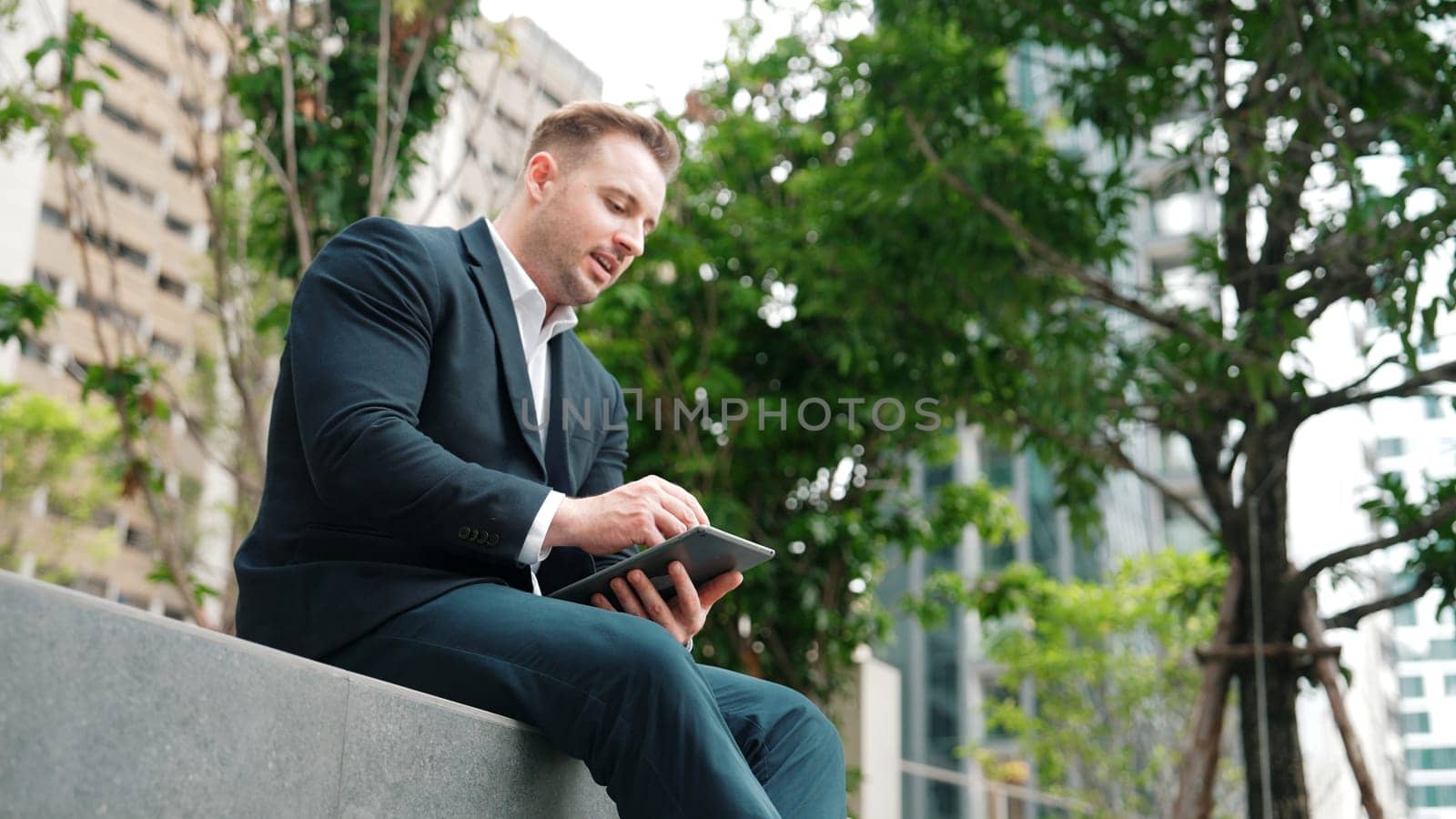 Professional businessman sitting at park while working on phone by reading report from marketing team. Happy manager looking increasing sales or successful project while smiling at eco city. Urbane