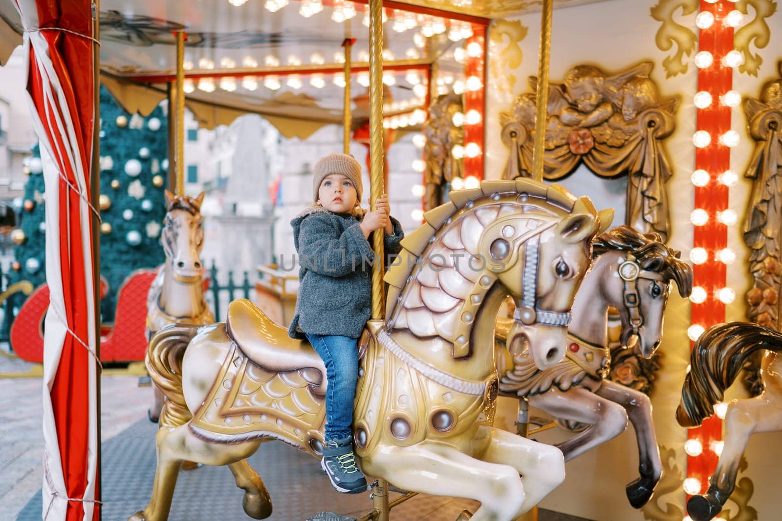 Little girl rides a toy horse on a carousel in the square near a decorated Christmas tree. High quality photo