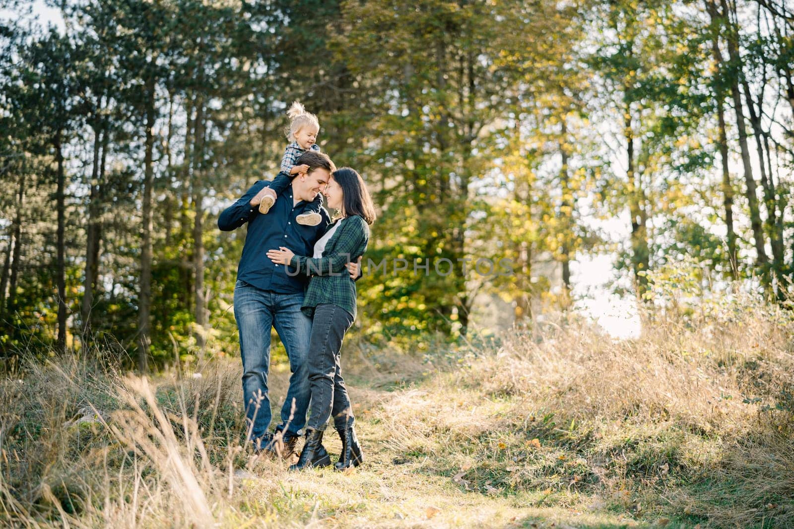 Mom hugs dad with a little girl on his shoulders touching his forehead with her forehead. High quality photo