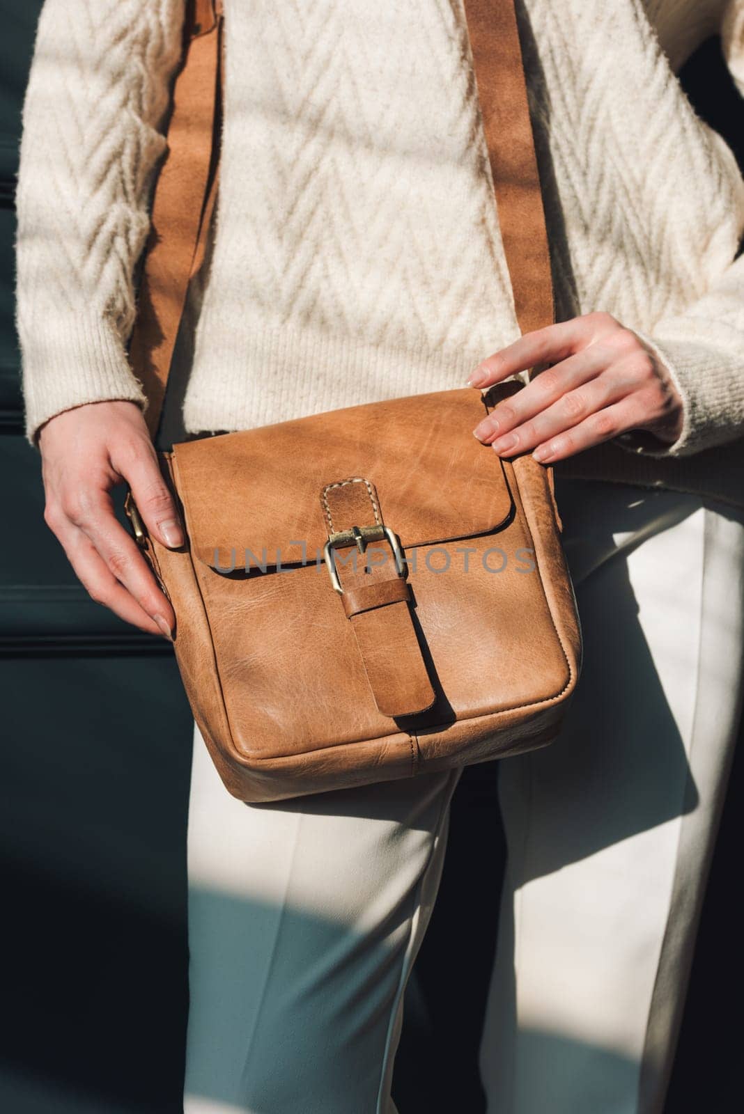 beautiful woman posing with a small brown bag near grey wall. Model wearing stylish white sweater and classic trousers