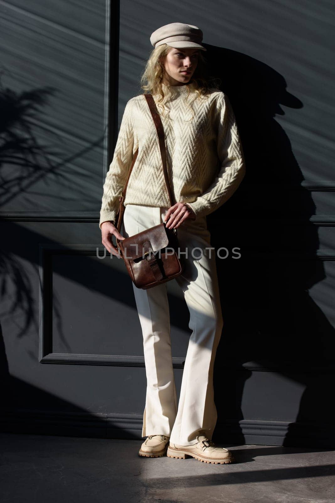 Studio portrait of beautiful woman with a curly blond hair holding black bag, posing on gray background. Model wearing stylish cap, sweater and classic trousers