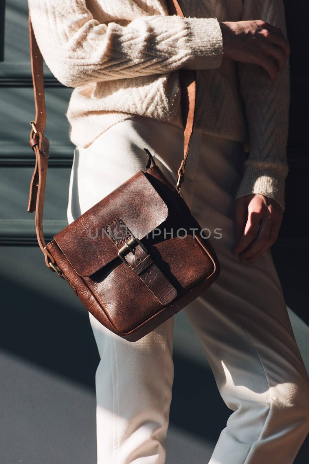 beautiful woman posing with a small brown bag near grey wall. Model wearing stylish white sweater and classic trousers