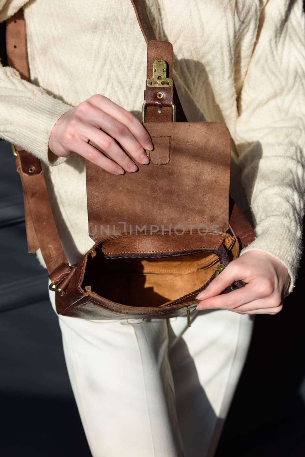 beautiful woman posing with a small brown bag near grey wall. Model wearing stylish white sweater and classic trousers