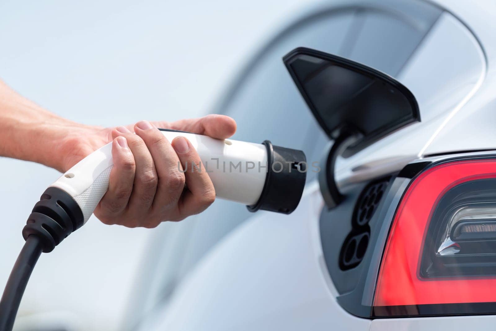 Hand insert EV charger plug into electric vehicle to recharge EV car battery from outdoor charging station. Fresh daylight environment with alternative clean and sustainable energy concept. Perpetual