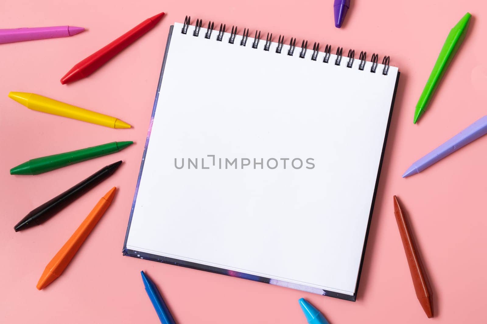 Open sketchbook with white empty blank. Colorful wax crayons on the pastel pink background. Flat lay composition