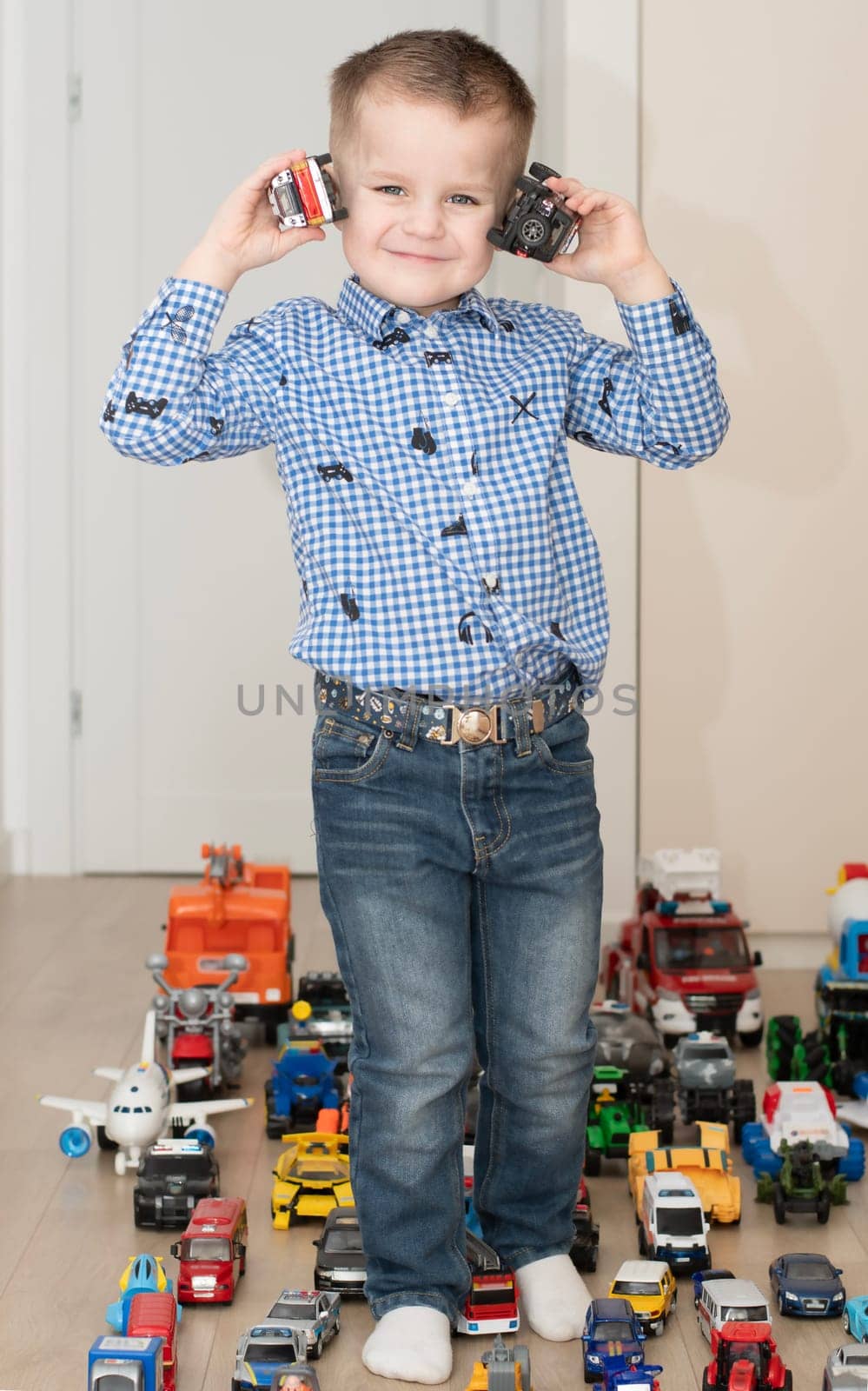Children. Toy cars. A small cheerful and handsome boy, 4 years old, wearing a checkered shirt, plays with many colorful cars in a home interior. The toys are placed evenly on the floor.