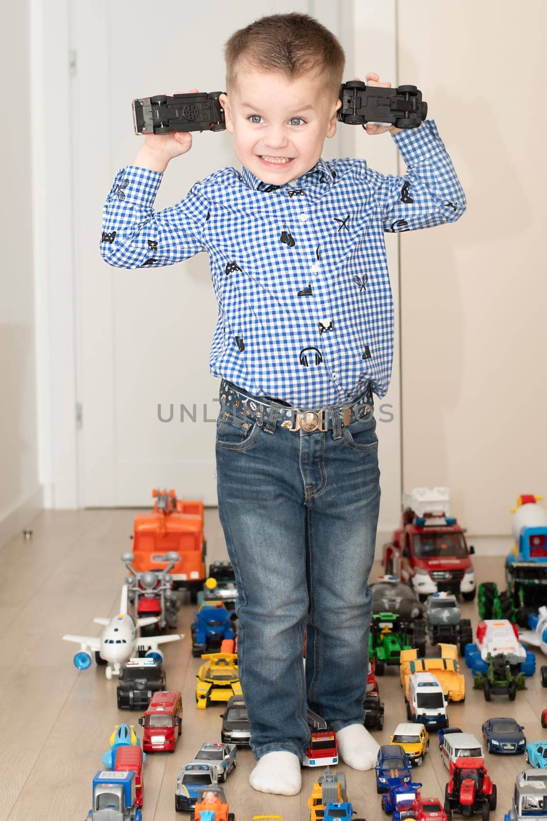 Children. Toy cars. A small cheerful and handsome boy, 4 years old, wearing a checkered shirt, plays with many colorful cars in a home interior. The toys are placed evenly on the floor.