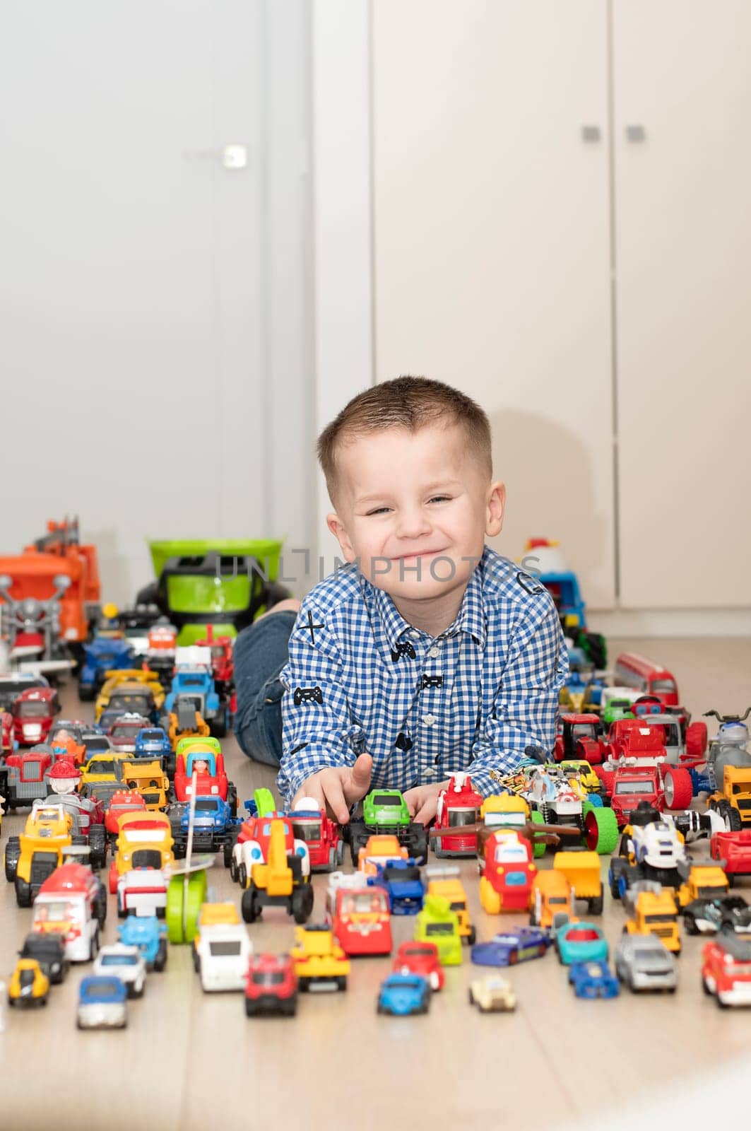 Concept of children's toys. A little boy, 4 years old, plays happily, lying on the floor, with colorful small and large cars in the children's room. Soft focus.