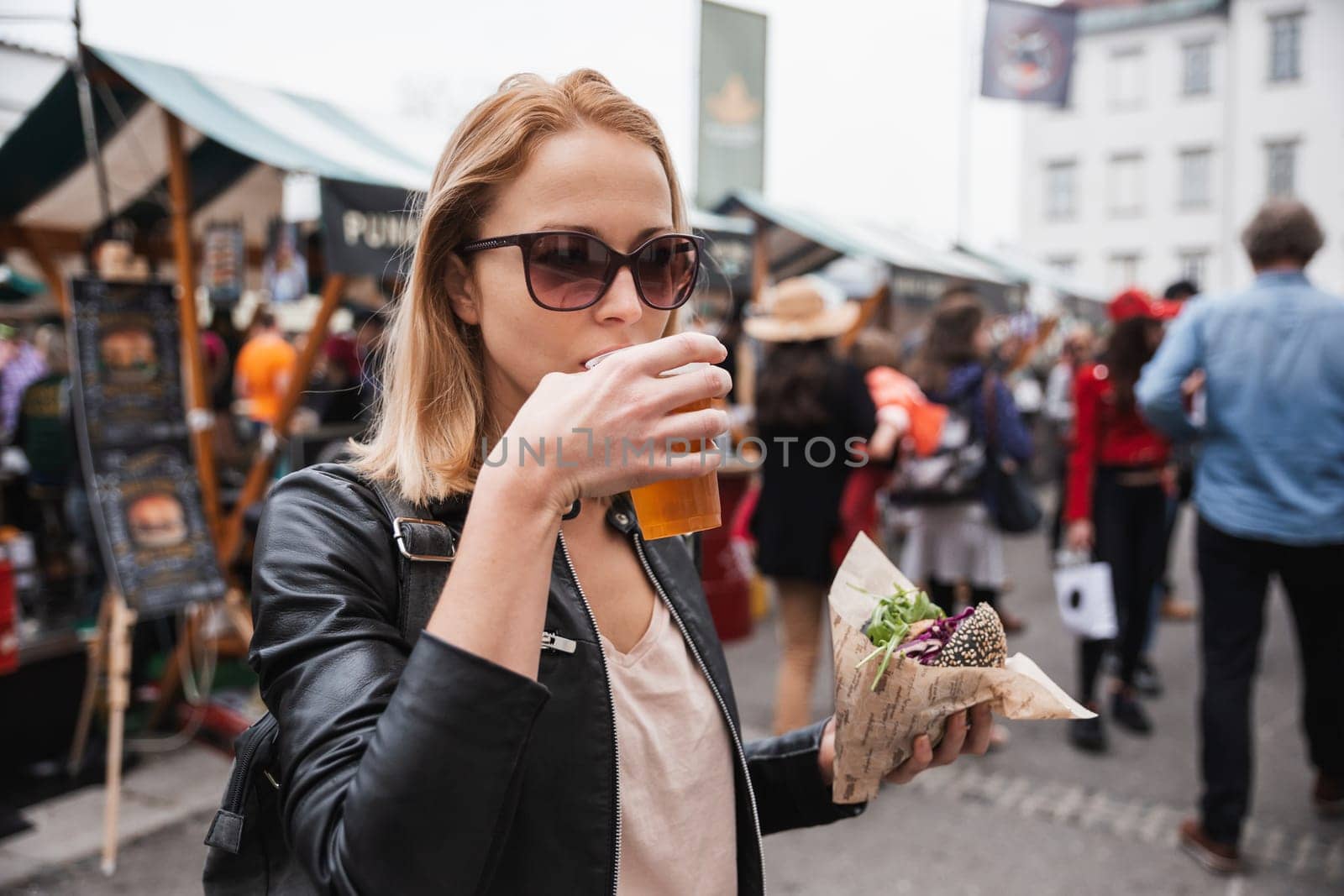 Beautiful young woman holding delicious organic salmon vegetarian burger and drinking homebrewed IPA beer on open air beer an burger urban street food festival in Ljubljana, Slovenia. by kasto