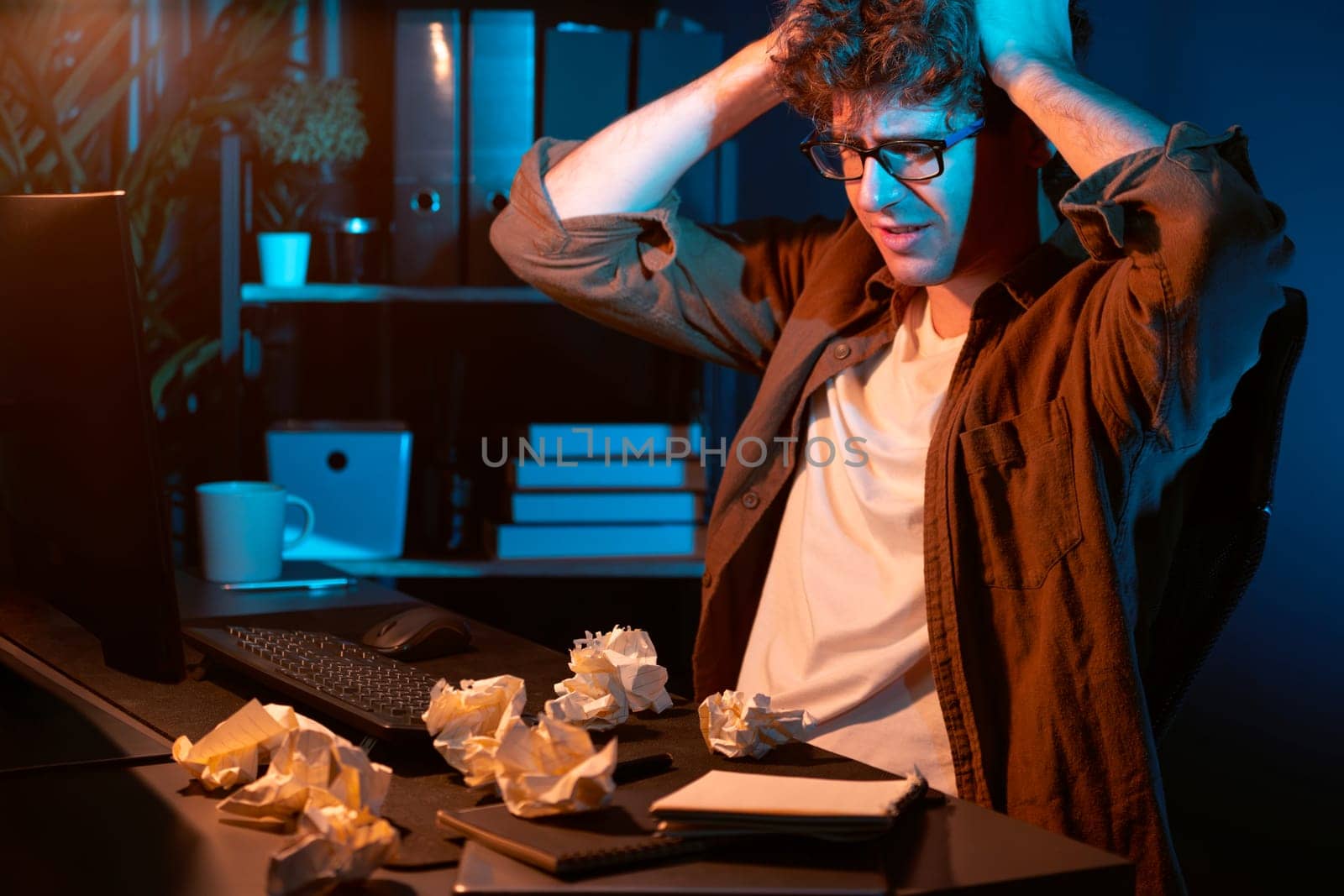Stressful creative manager working with overworked project surrounded crumpled papers while waiting email sending back on laptop screen in casual shirt at night time at neon modern office. Gusher.