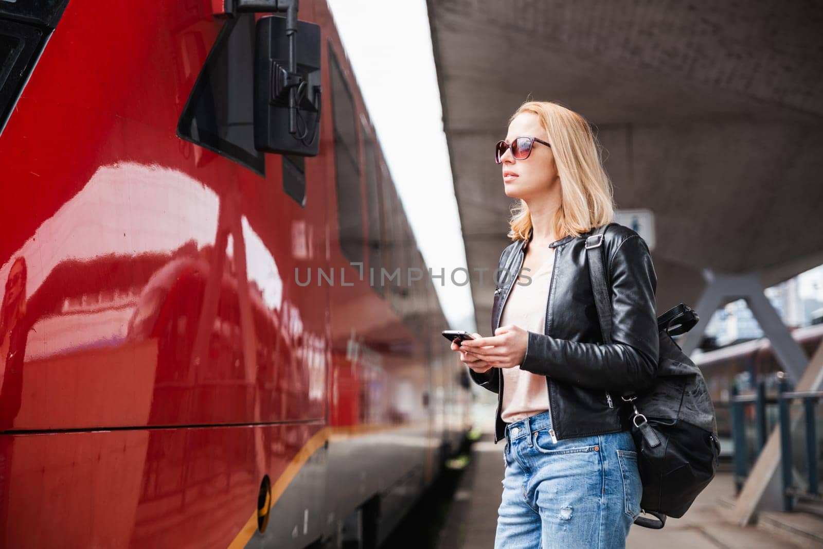 Young blond woman in jeans, shirt and leather jacket wearing bag and sunglass, embarking red modern speed train on train station platform. Travel and transportation