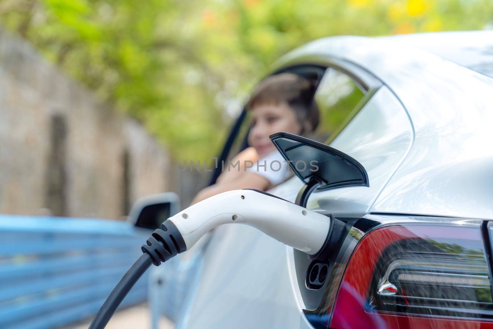 EV car recharge battery from EV charging station with blur little boy waiting inside the car in background. Road trip travel by alternative vehicle powered by clean and sustainable energy. Perpetual