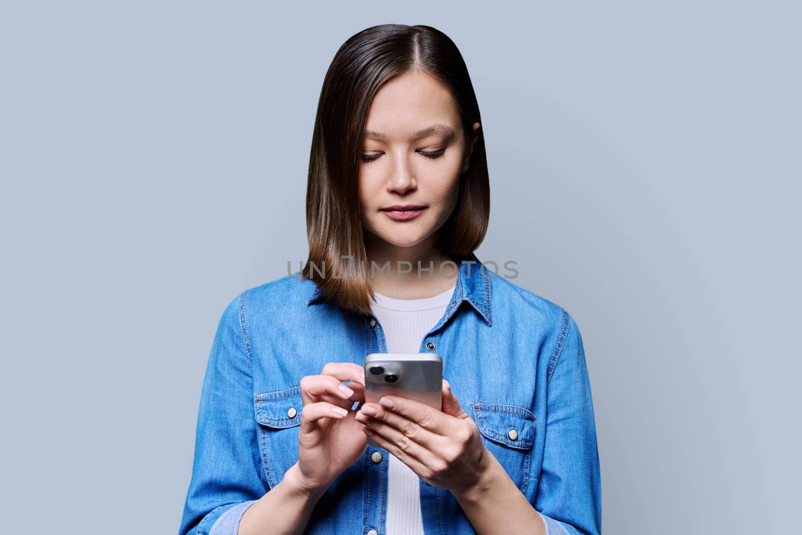 Young woman using smartphone in gray background. Serious 20s female looking at screen, texting. Mobile Internet applications apps, technologies for work education communication shopping healthcare