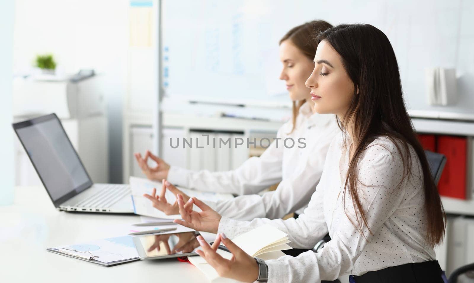 Portrait of gorgeous businesswoman closing tender eyes in calmness and satisfaction sitting at modern workplace. Beautiful woman doing special meditation pose. Friendly teamwork concept