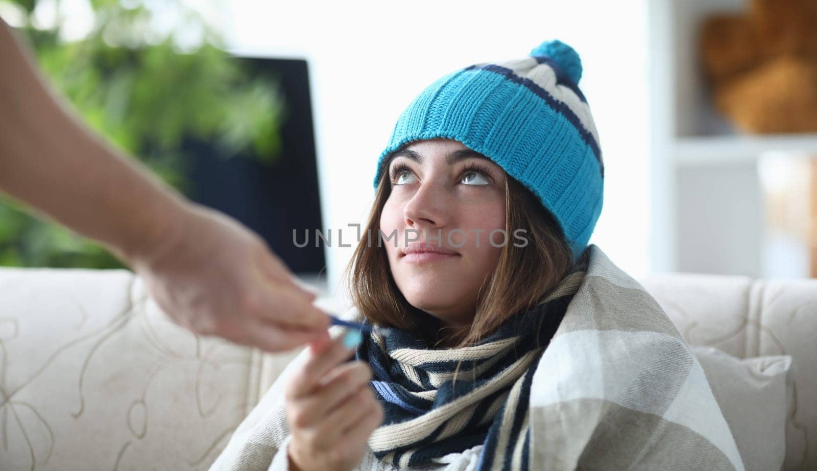 Young sick woman in hat and scarf sits under blanket on sofa at home and receives medicine. Outpatient pneumonia treatment concept.