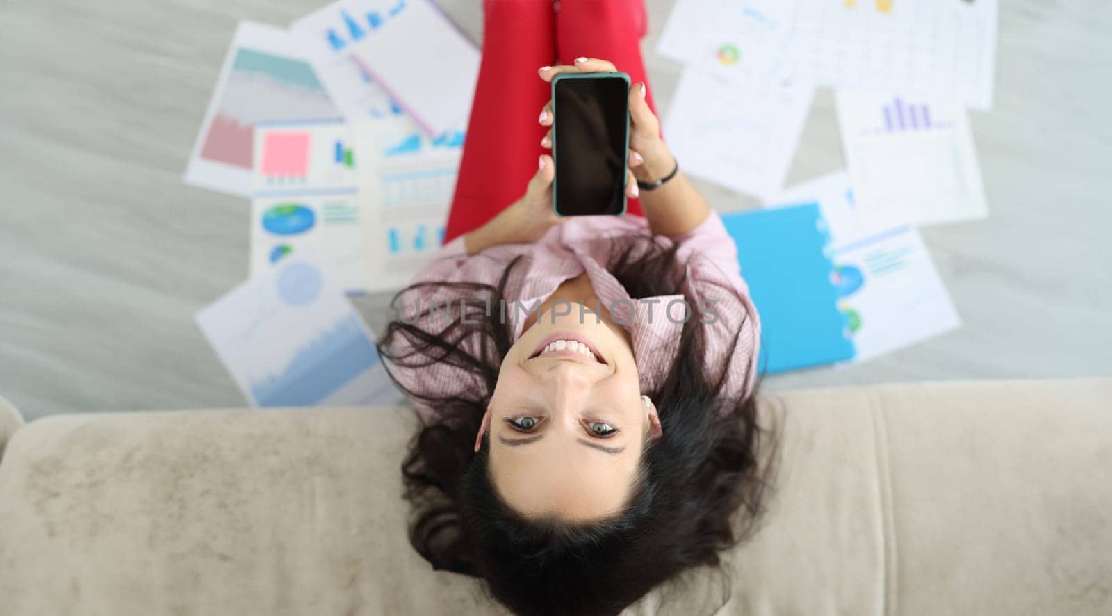 Happy woman sit on floor put her head on sofa. Female hand hold phone screen up. On floor many document with diagrams top view.