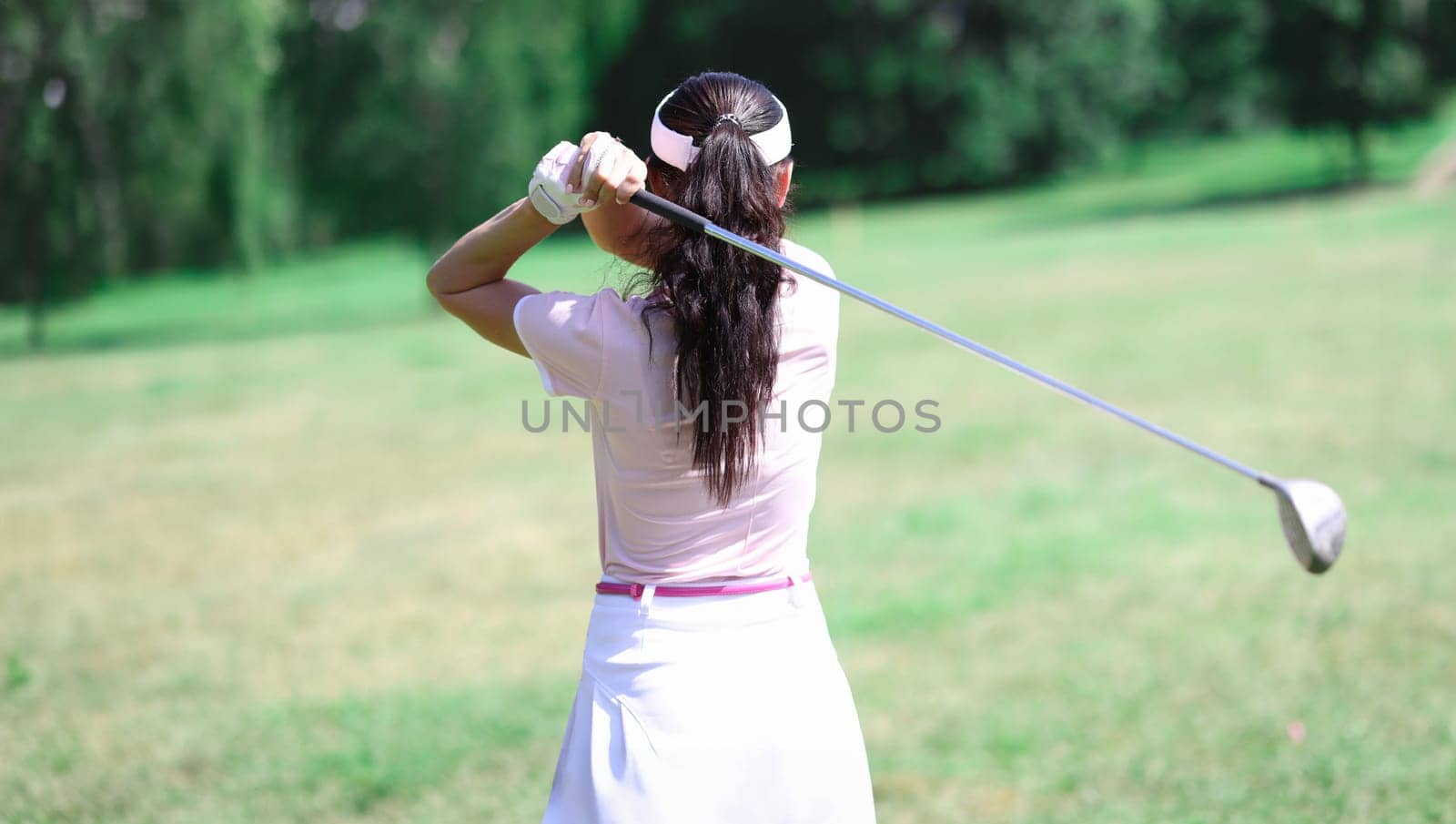Woman in suit hold golf club behind her back. Back view of person playing golf on green lawn.